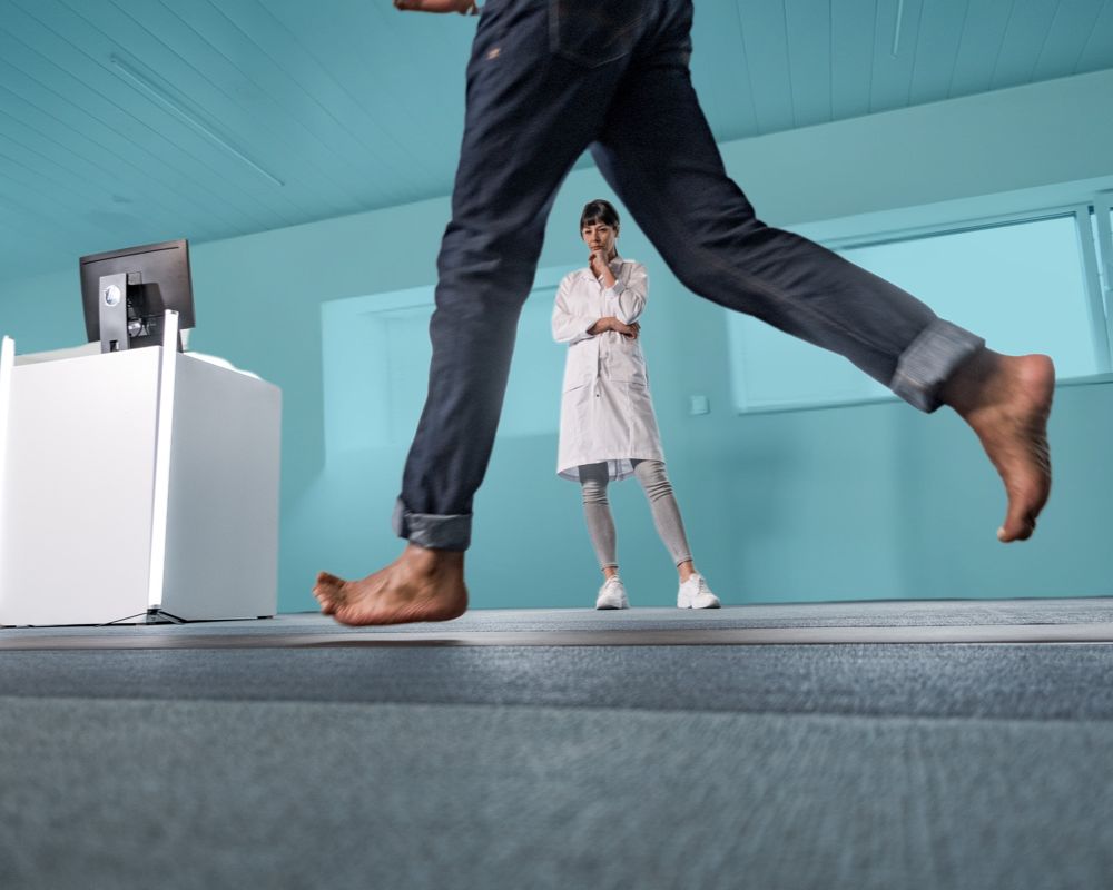 Gait analysis assessment: man wearing jeans running barefoot on a Footscan pressure pad while a physiotherapist in a white coat looks on