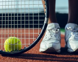 cropped image of a person wearing tennis shoes with a racquet and ball on a tennis court: image showing how gait analysis can benefit anyone playing tennis in Helensburgh