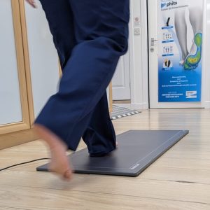 Physiotherapist demonstrates what hppens in a Footscan advanced gait analysis appointment by walking on a pressure mat on a wooden floor at The Lomond Clinic