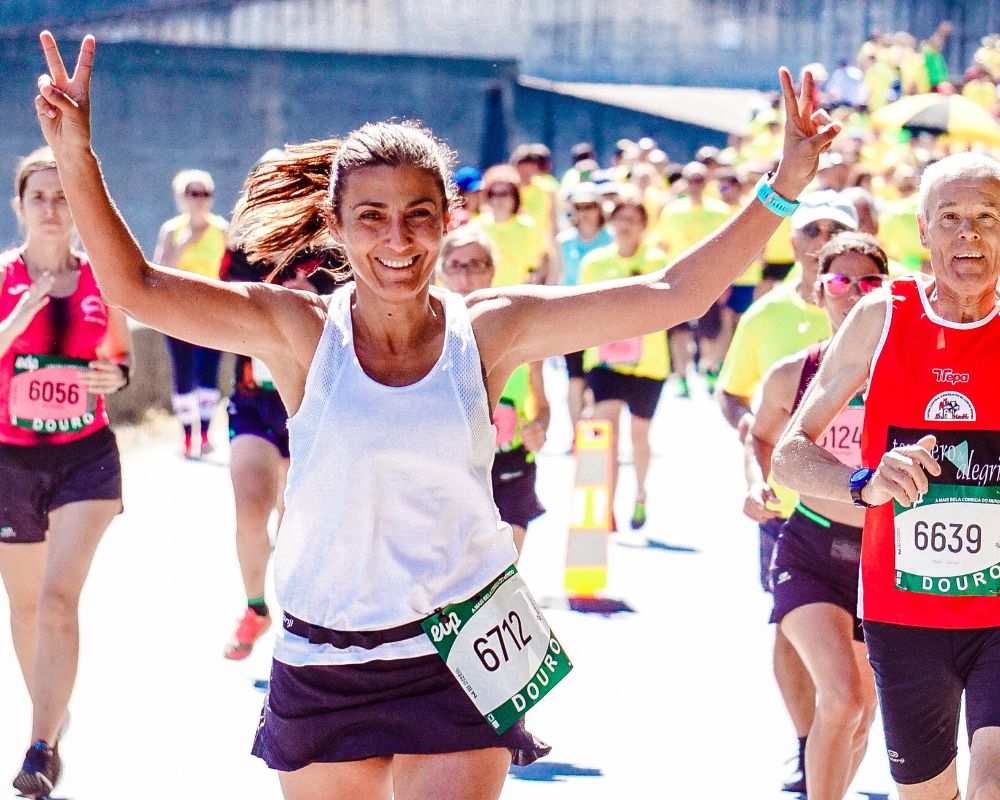 Female runner wearing white vest, navy shorts and a race number raises her arms and smiles boardly at the camera amongst competitors in a road race