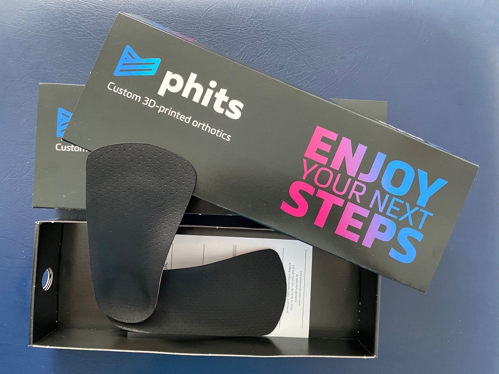 Phits custom 3D-printed orthotics lying inside black branded packaing featuring the Phits logo in blue and white and pink and blue graduated lettering saying 'Enjoy your next steps'