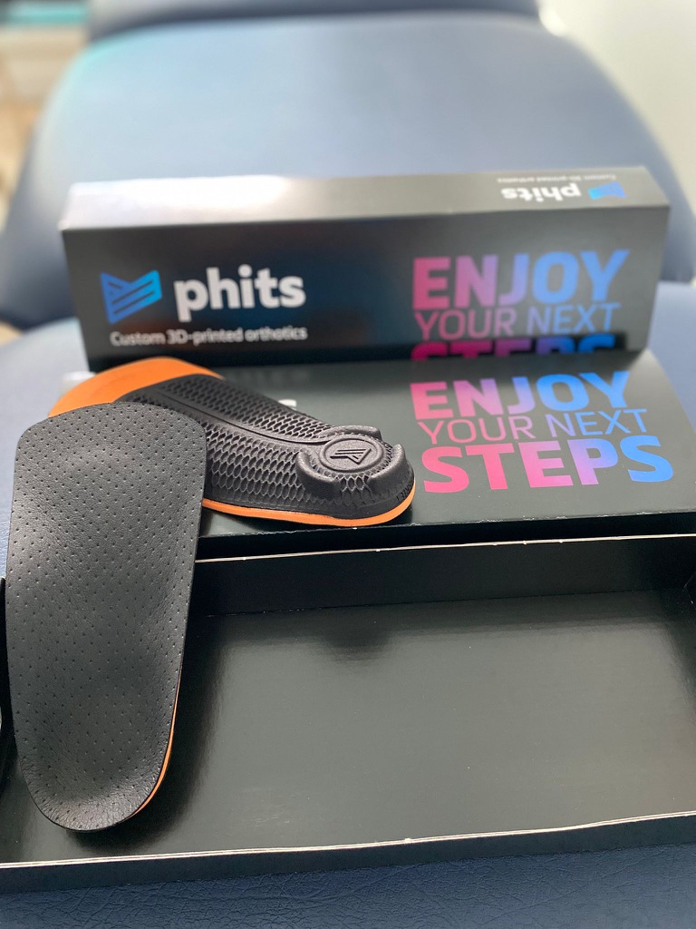 Photo of Phits custom 3d printed othoses (insoles) and their packaging, a black box printed with the Phits logo and 'Enjoy your next steps' by The Lomond Clinic, Helensburgh