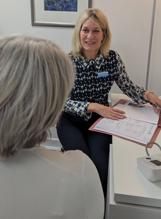 Dr Joyce Braaksma smiles at a female patient during a consultation at a white desk with patient notes and a blood pressure monitor.