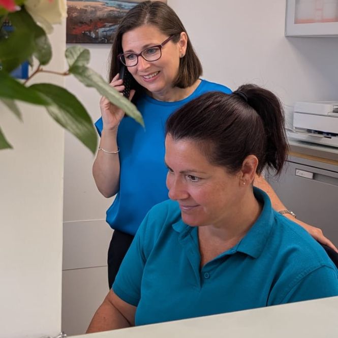 Receptionists Joanne and Rachael at The Lomond Clinic reception desk. Joanne wears glasses and stands on the phone. Rachael sits beside her. Both look at a computer screen out of shot.